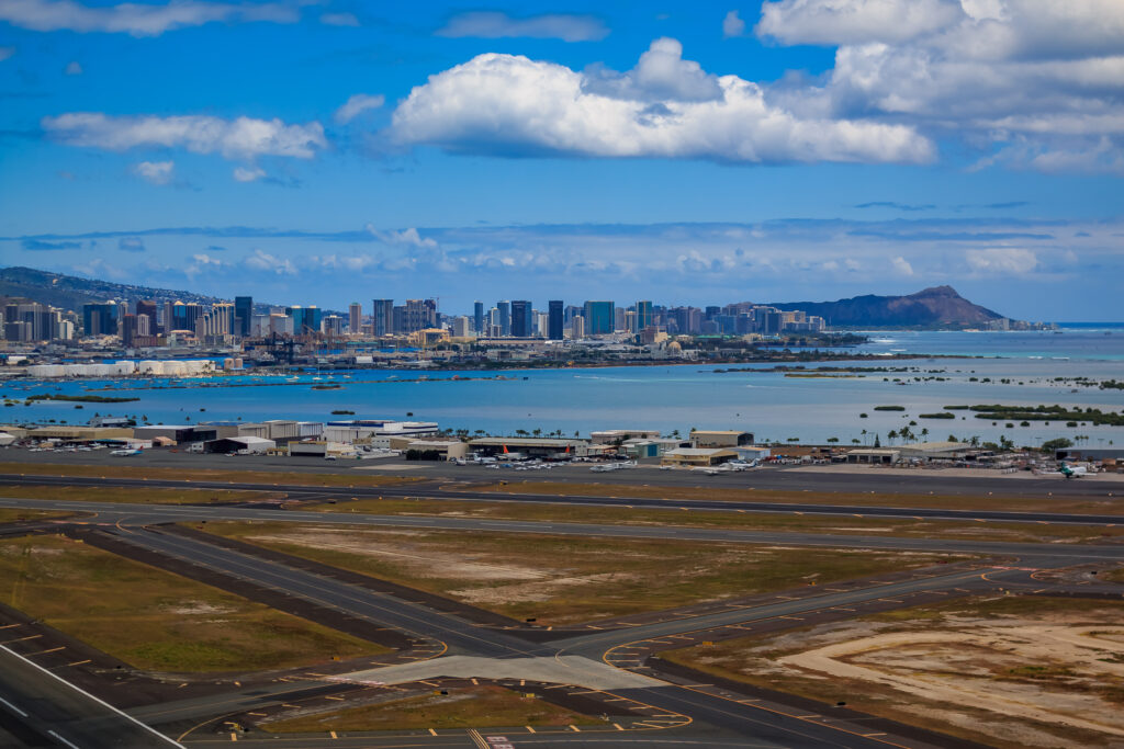 Aerial view of an airport runway with a coastal city skyline, tall buildings, and mountains in the background under a partly cloudy sky.