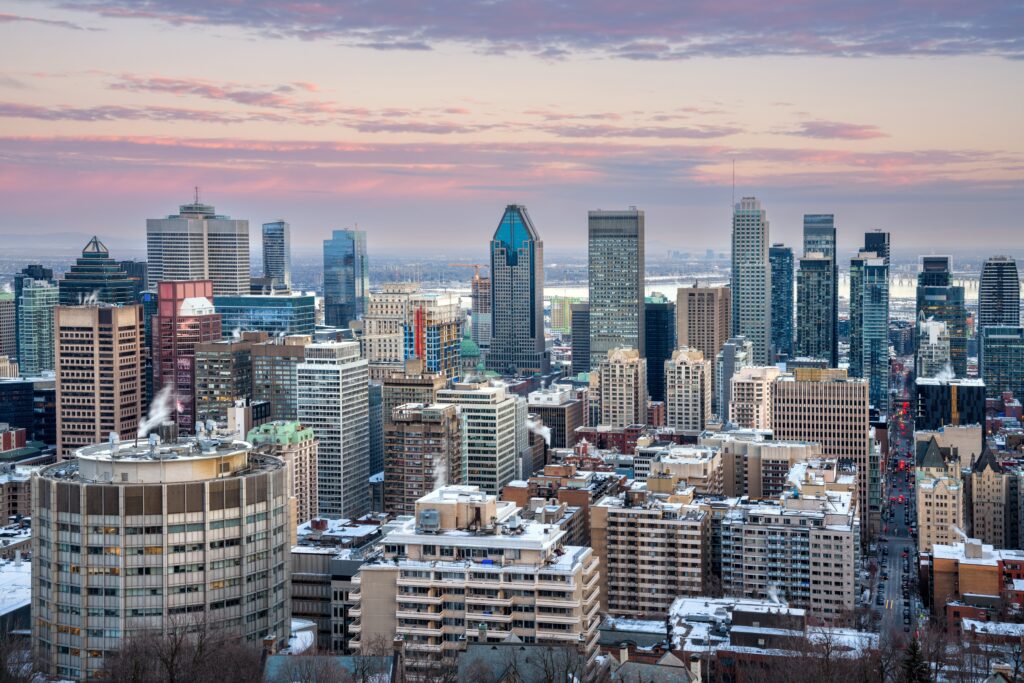 Downtown Montreal skyline at sunset, with tall modern skyscrapers, older buildings in the foreground, and patches of snow on rooftops under a pink and blue sky.