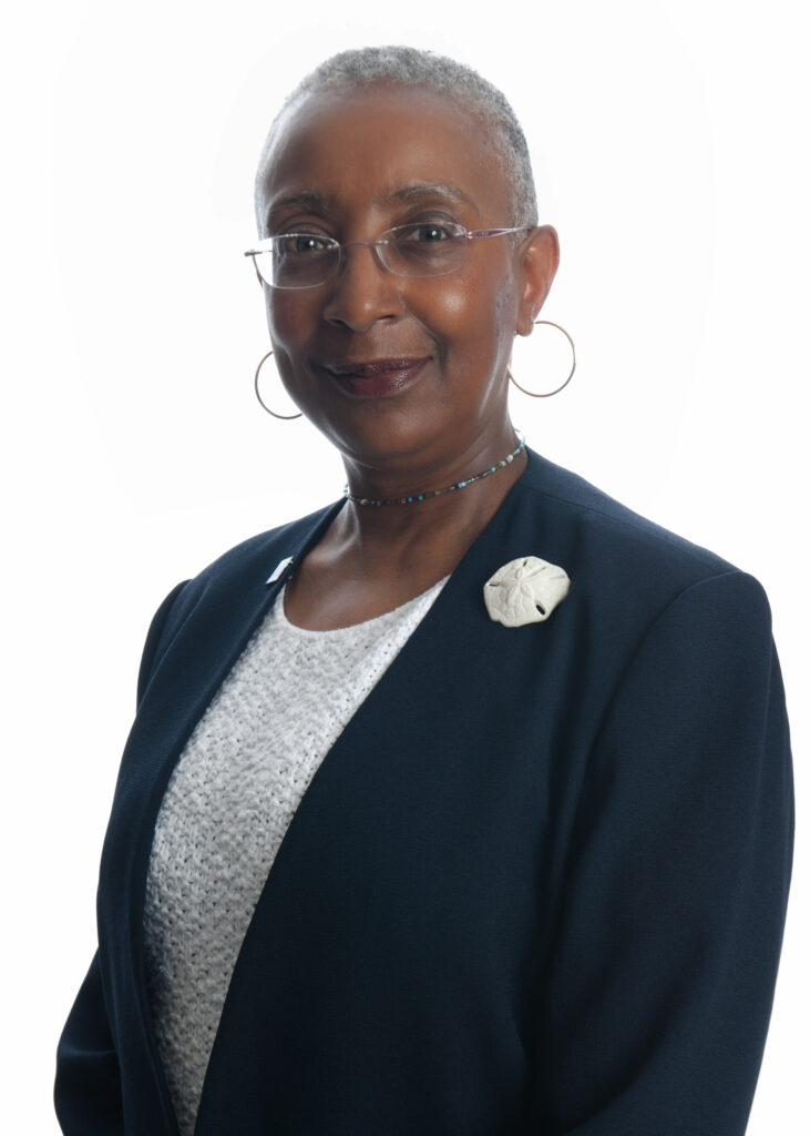 A woman with short gray hair, glasses, and hoop earrings, wearing a navy blazer with a white blouse and flower brooch, smiles slightly while standing against a plain white background.