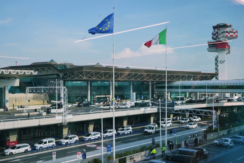 A view of an airport terminal with the European Union and Italian flags flying outside, several white taxis parked in front, and a control tower visible in the background under a clear blue sky.