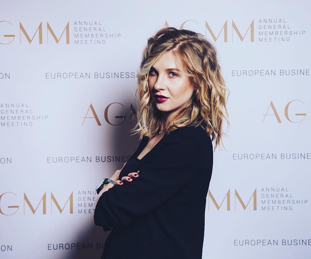 A woman with wavy blonde hair, wearing a black jacket, stands with arms crossed and looks at the camera in front of a step-and-repeat banner that reads AGMM Annual General Membership Meeting.