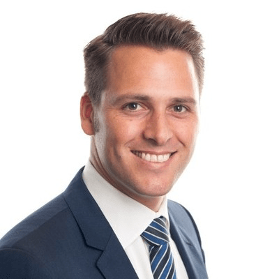 A man in a blue suit, white shirt, and striped tie is smiling at the camera against a plain white background. He has short brown hair styled neatly and light skin.