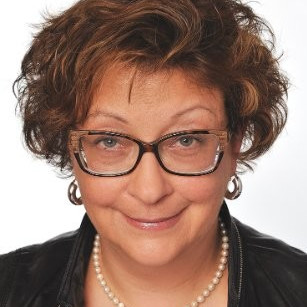 A woman with short, curly brown hair wearing glasses, hoop earrings, a pearl necklace, and a black leather jacket smiles at the camera against a white background.