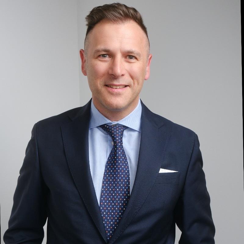 A man in a navy blue suit, light blue shirt, and patterned tie smiles while standing against a plain light gray background.