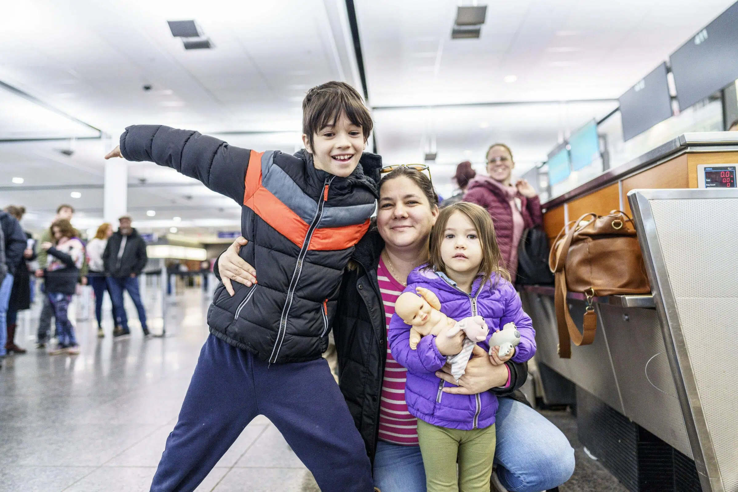 A woman kneels in an airport with a smiling boy in a black and orange jacket on her left and a girl holding two dolls on her right. Travelers and luggage are visible in the background.