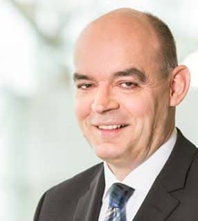 A middle-aged man with a bald head, wearing a dark suit, white shirt, and striped tie, smiling at the camera against a blurred light background.