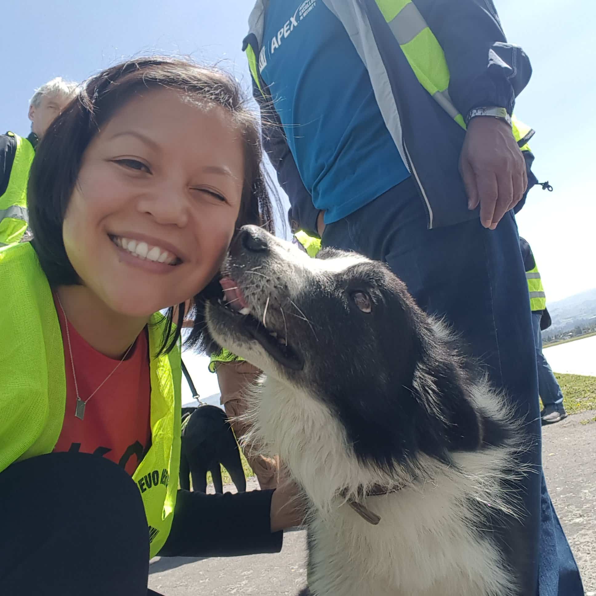 A smiling person in a yellow safety vest poses closely with a black and white dog outdoors. The dog looks up at the person affectionately. Other people in safety vests are visible in the background.