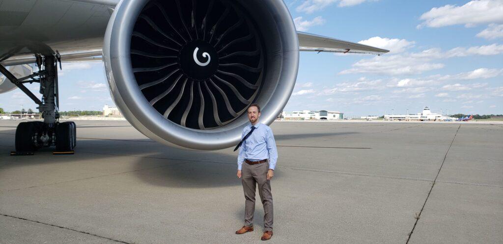 A man in business attire stands on an airport tarmac near the large engine of a commercial airplane, with airport buildings and a blue sky with scattered clouds in the background.