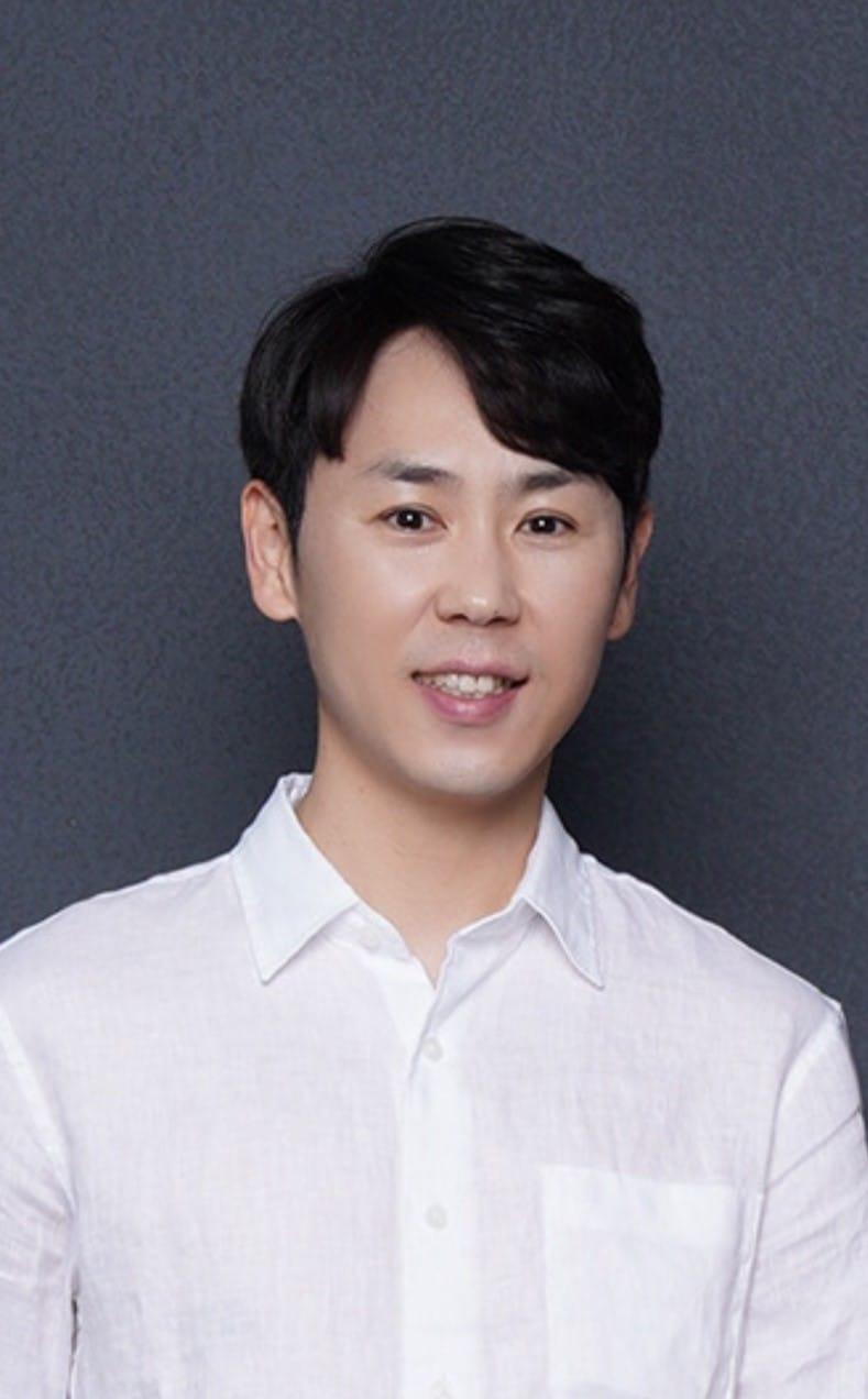 A man with short black hair is smiling at the camera, wearing a white collared shirt, standing against a plain dark gray background.