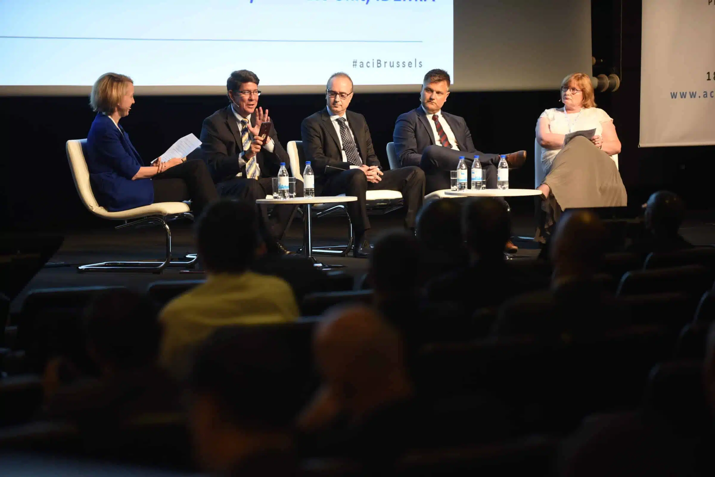 Five people in business attire sit on a stage panel discussion, with microphones and water bottles on a table. An audience watches, and a large screen is behind the speakers.