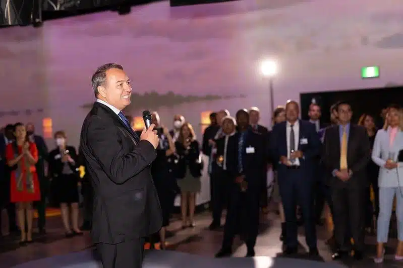 A man in a suit speaks into a microphone on stage at a formal event, while a group of people in business attire watch and listen attentively in a softly lit room.