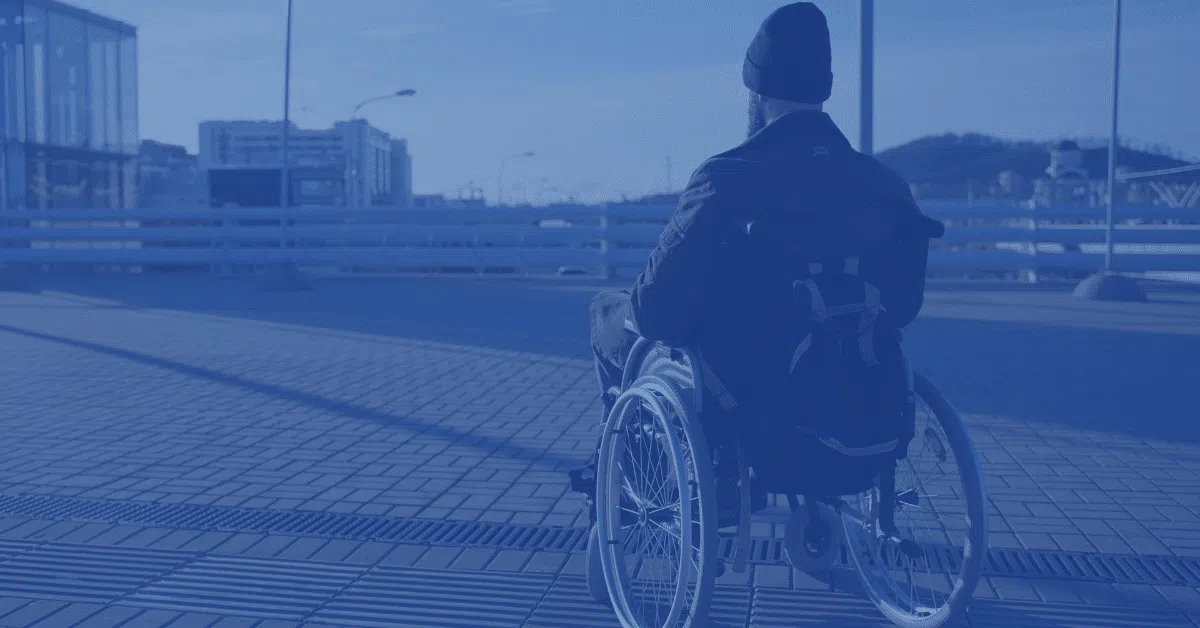 A person in a wheelchair faces away from the camera, outdoors on a paved area near buildings and streetlights, wearing a beanie and backpack, with a blue tint over the image.