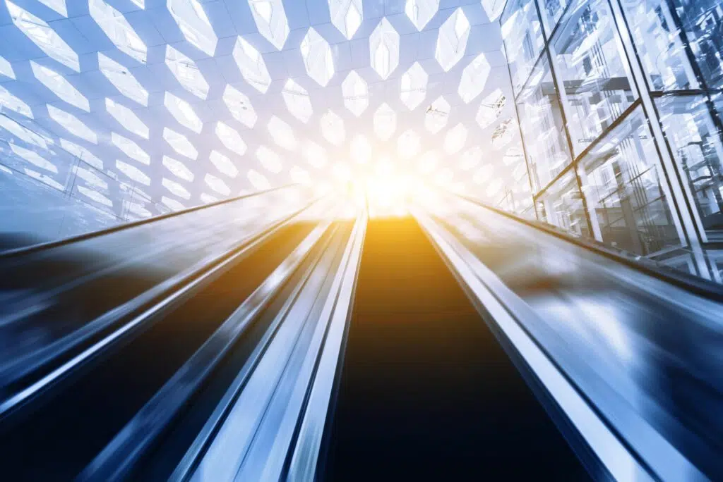 A blurred view looking up an escalator toward bright sunlight, with a geometric glass ceiling and modern architecture visible above.