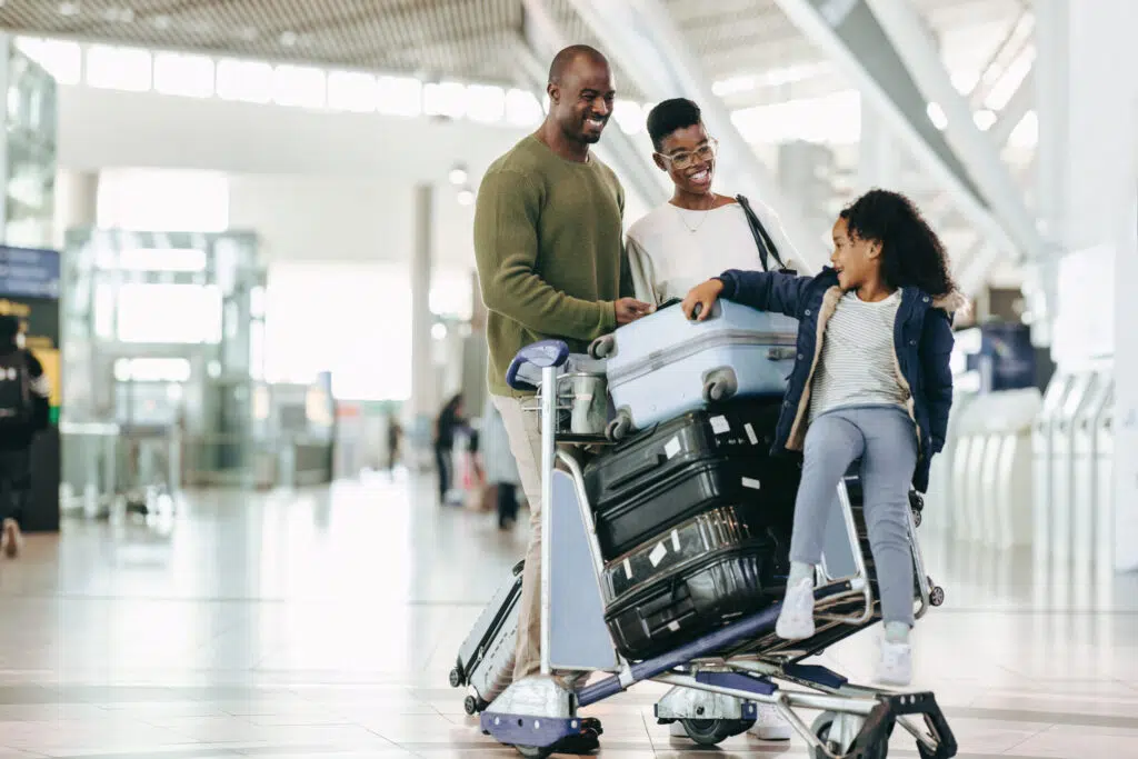 A smiling family with two adults and one child in an airport, standing by a luggage cart stacked with suitcases. The child sits playfully on the cart while the adults look at her happily.