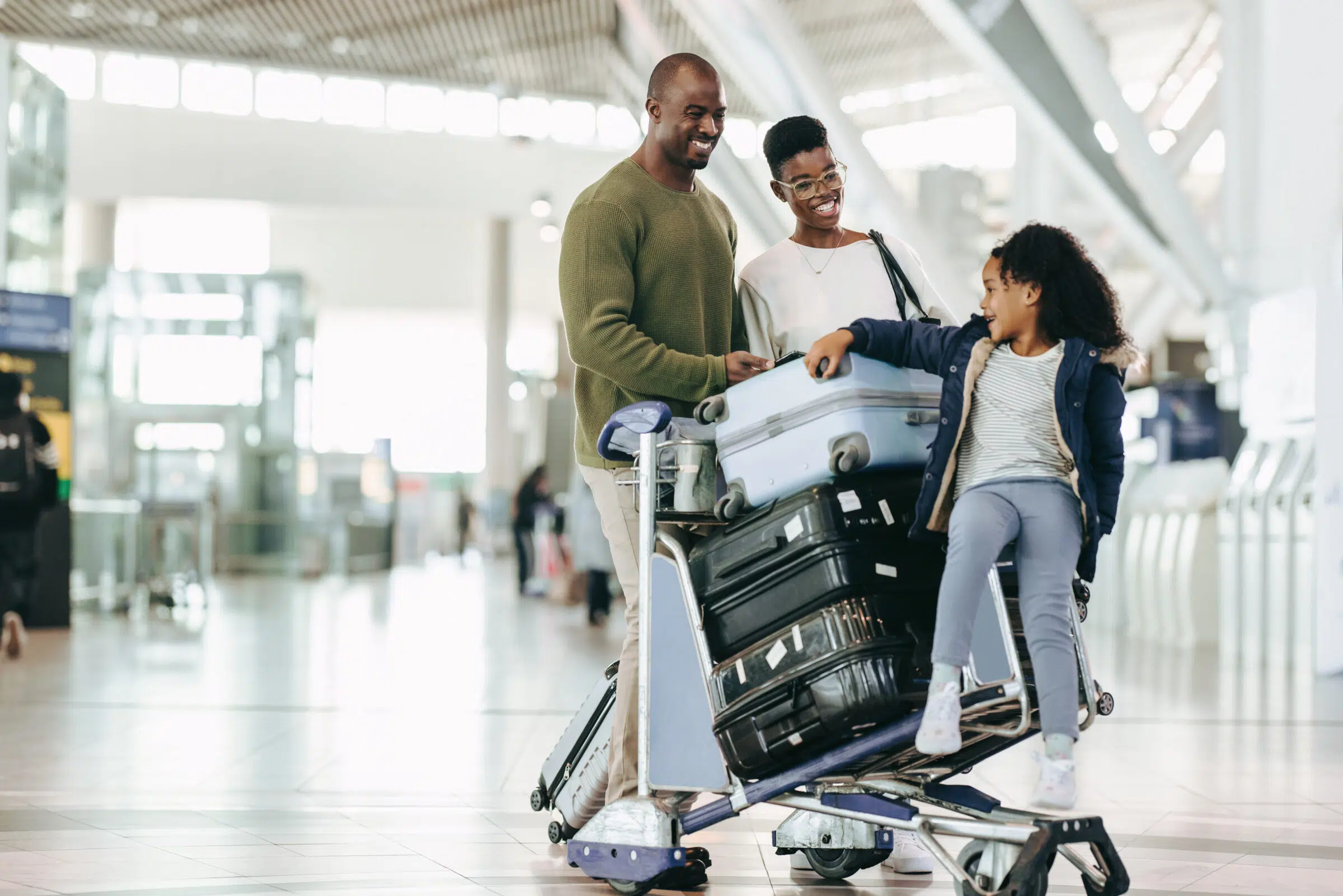 A smiling family with two adults and one child in an airport, standing by a luggage cart stacked with suitcases. The child sits playfully on the cart while the adults look at her happily.