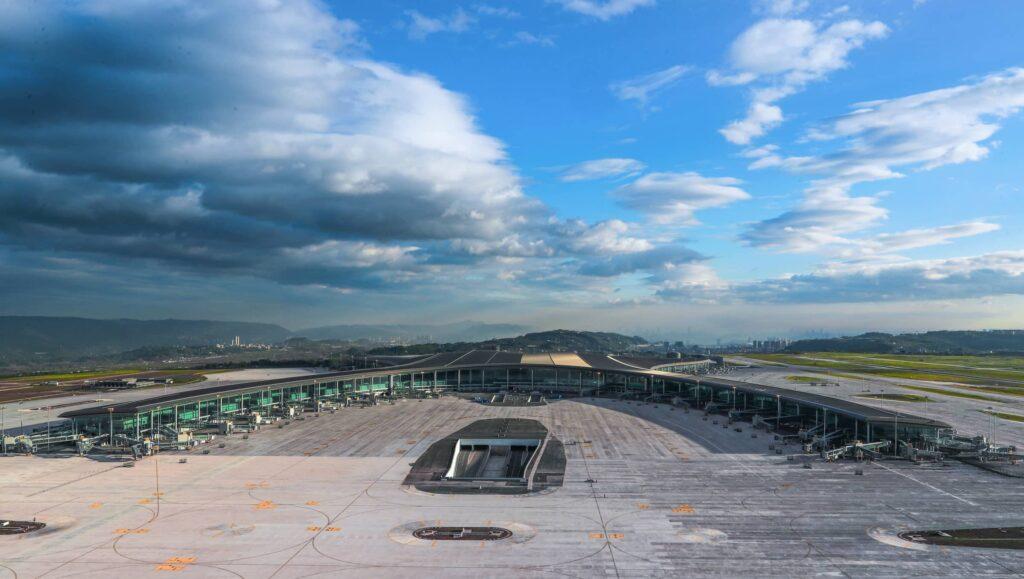 A wide view of a modern airport terminal with multiple gates, empty tarmac, and surrounding runways under a partly cloudy blue sky, with distant hills in the background.