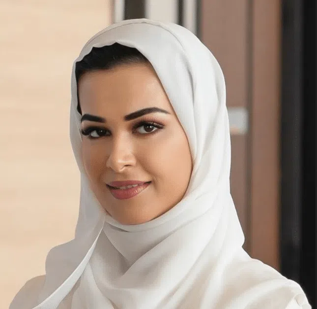 A woman wearing a white hijab and a white top smiles softly at the camera in an indoor setting with neutral-colored walls.