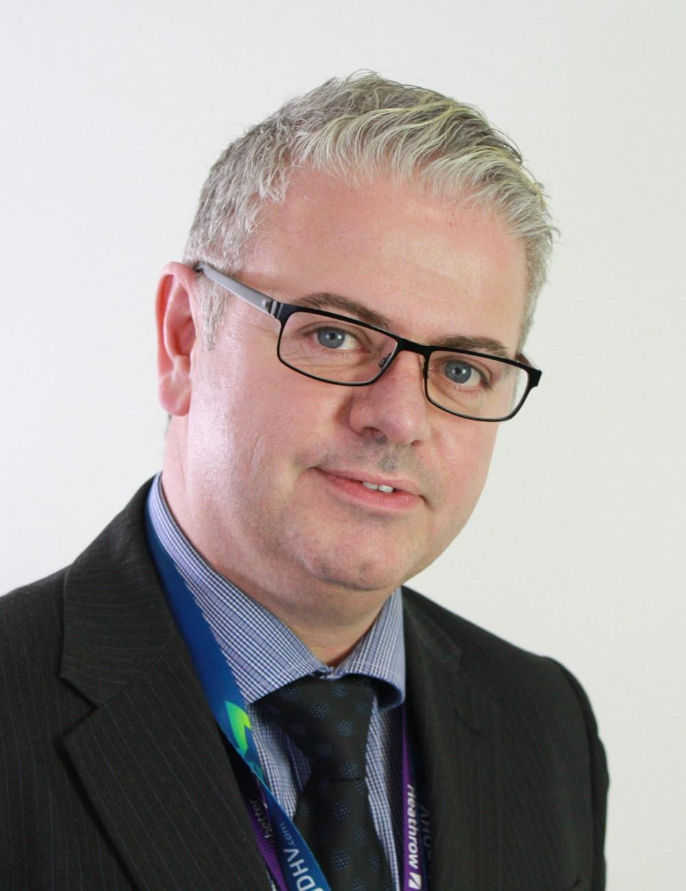 A man with short gray hair and glasses wearing a dark pinstripe suit, blue shirt, and dark tie, poses against a plain light background.