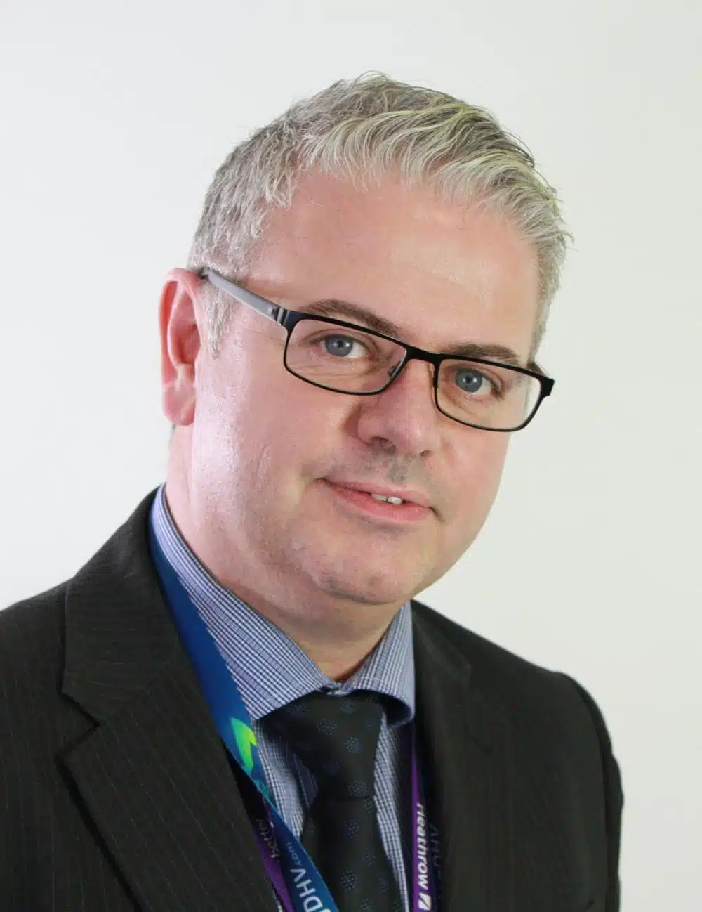A man with short gray hair and glasses wearing a dark pinstripe suit, blue shirt, and dark tie, poses against a plain light background.