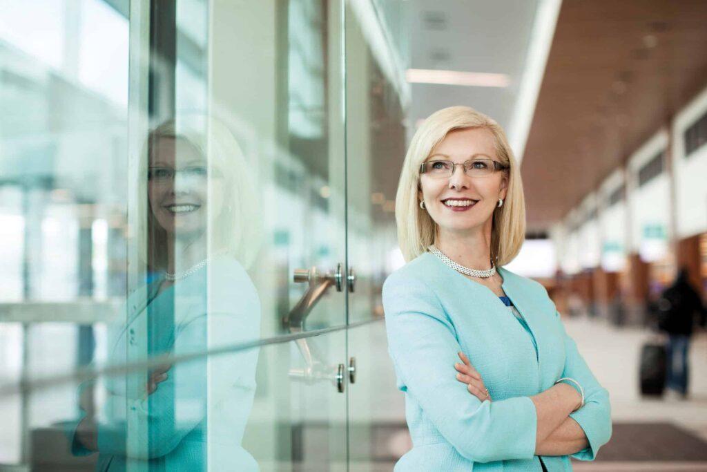 A smiling woman with blonde hair and glasses stands confidently with arms crossed, wearing a light blue suit and pearl necklace, in a modern hallway with glass walls reflecting her image.