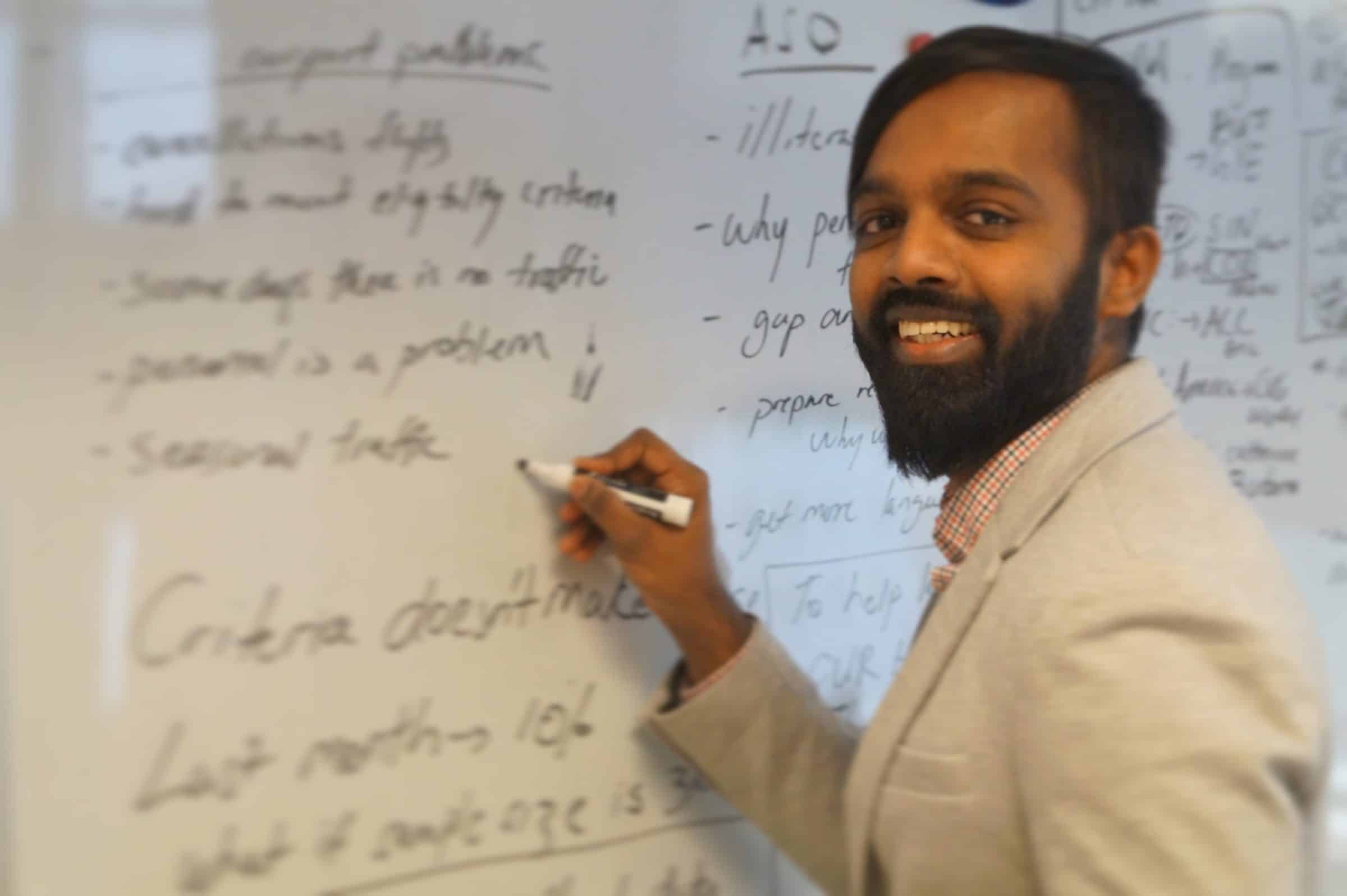 A man in a light-colored blazer smiles at the camera while writing on a whiteboard filled with notes and diagrams.
