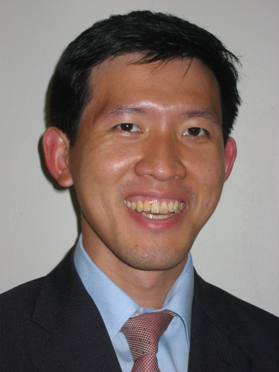 A man with short black hair smiles at the camera. He is wearing a dark suit jacket, a light blue dress shirt, and a patterned tie. The background is plain and light-colored.