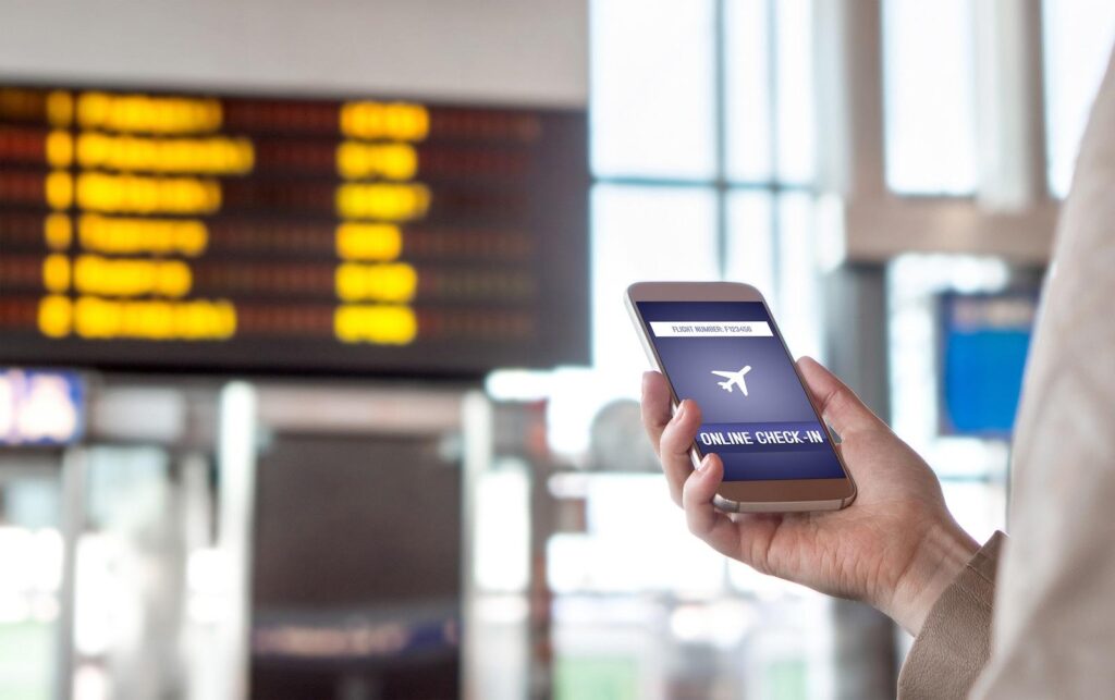 A person holding a smartphone with an online check-in screen at an airport, with a large flight information display board visible in the background.