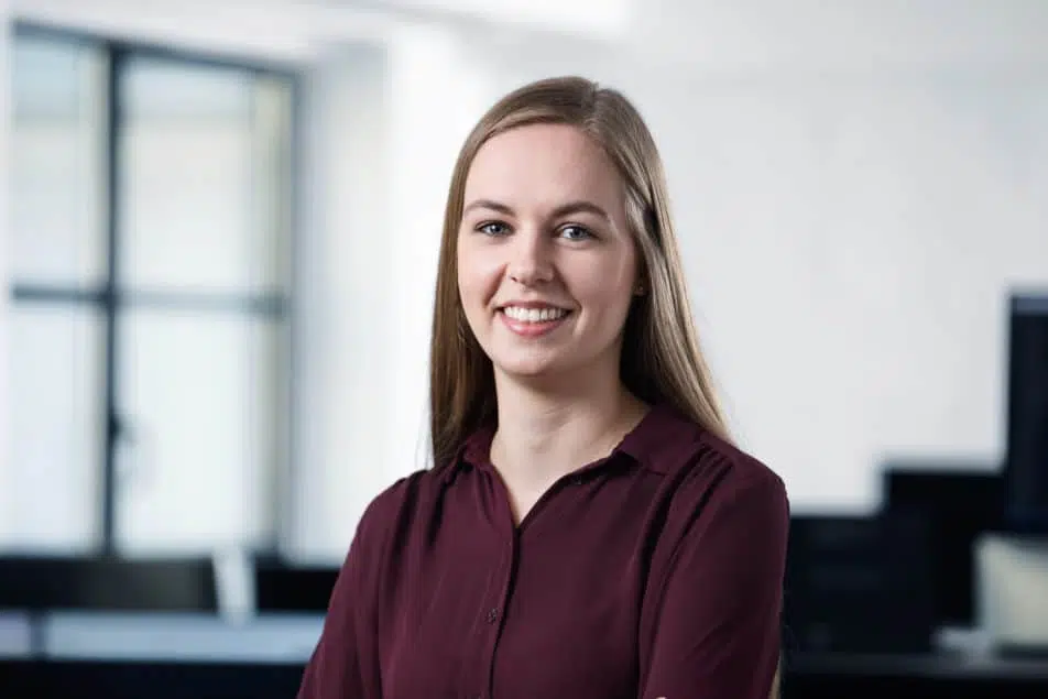 A young woman with long straight hair, wearing a maroon blouse, stands smiling in an office setting with blurred windows and computer screens in the background.