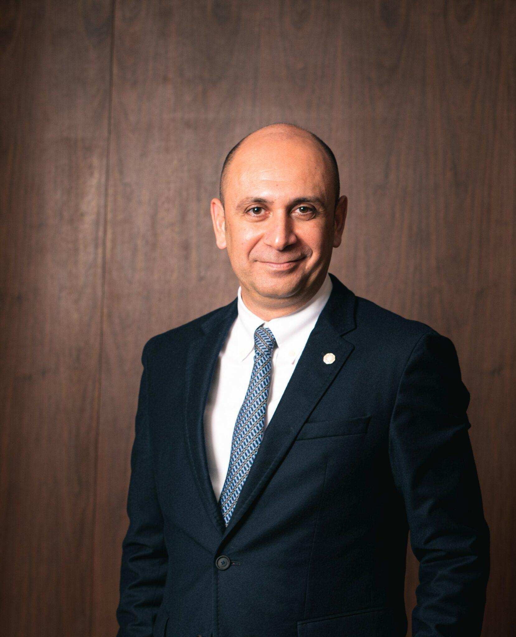 A man in a dark suit, white shirt, and patterned tie stands in front of a wooden background, smiling at the camera.