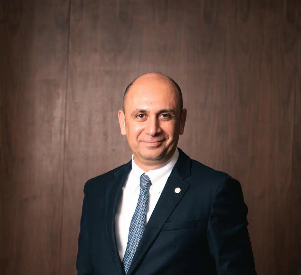 A man in a dark suit and patterned tie stands in front of a wooden wall, smiling slightly at the camera.