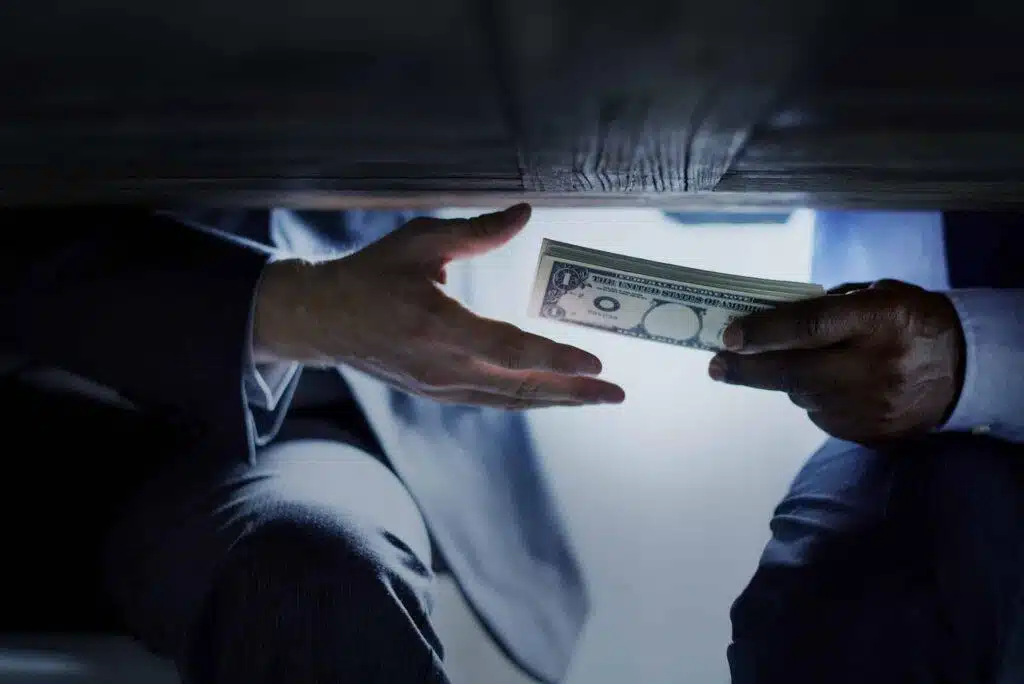 Two people in business suits exchanging a stack of US dollar bills under a table, suggesting a secretive or possibly illicit transaction. Only their hands, arms, and part of their legs are visible.