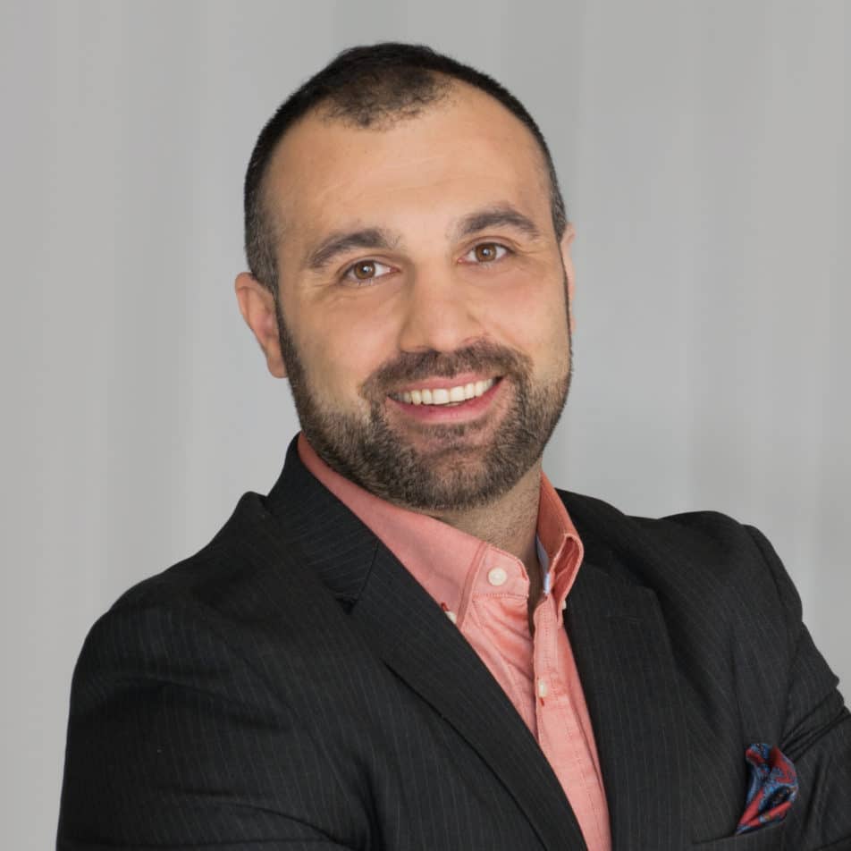 A man with short dark hair and a beard smiles confidently. He is wearing a black suit jacket over a pink shirt and stands with his arms crossed against a plain, light background.