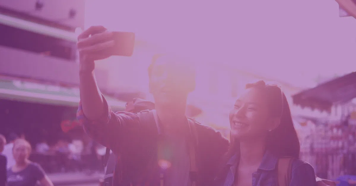 Two people smile and take a selfie together outdoors on a city street, with sunlight creating a bright pink filter over the image.