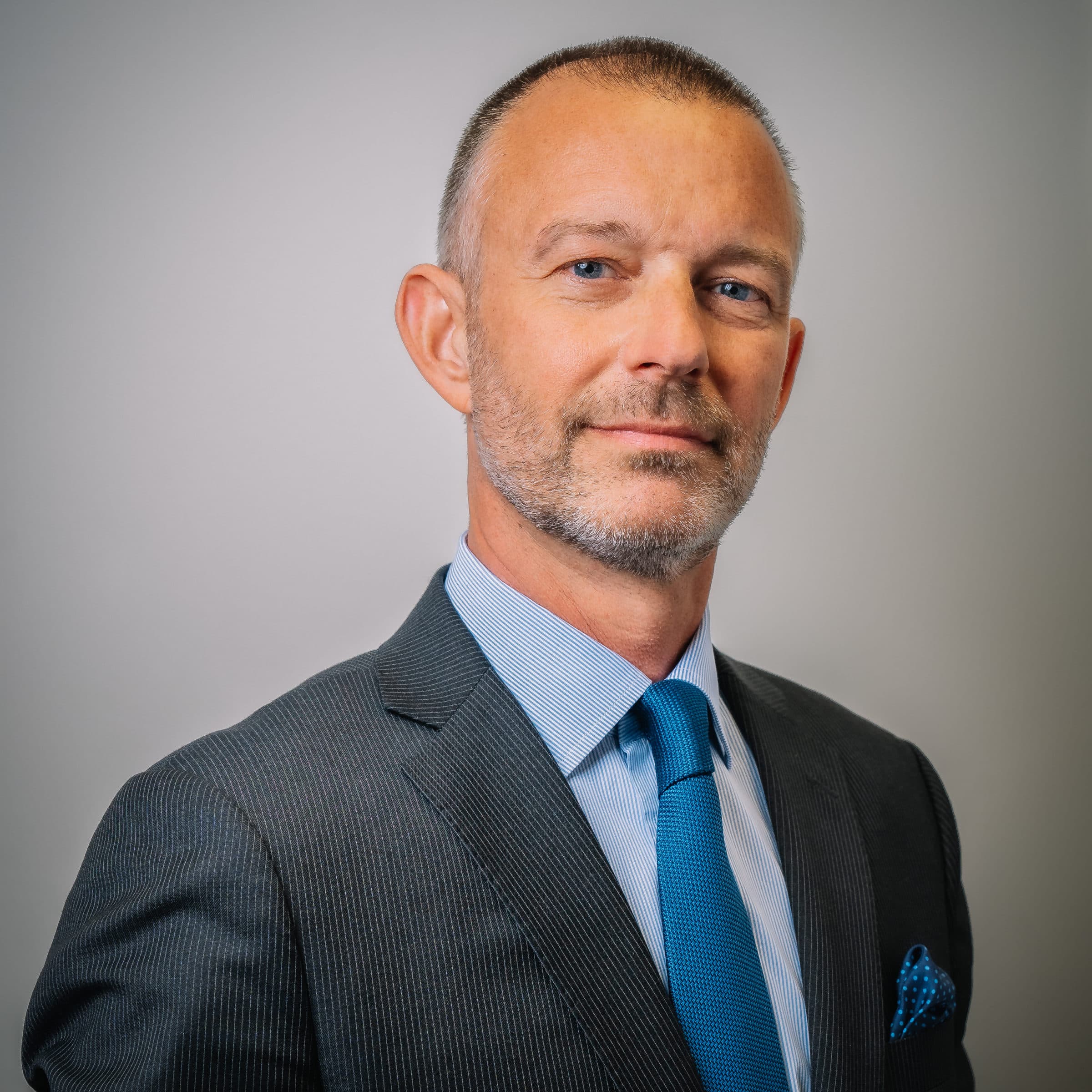 A confident middle-aged man with short hair and a trimmed beard, wearing a gray suit, light blue shirt, and blue tie, stands against a plain light background, looking at the camera.