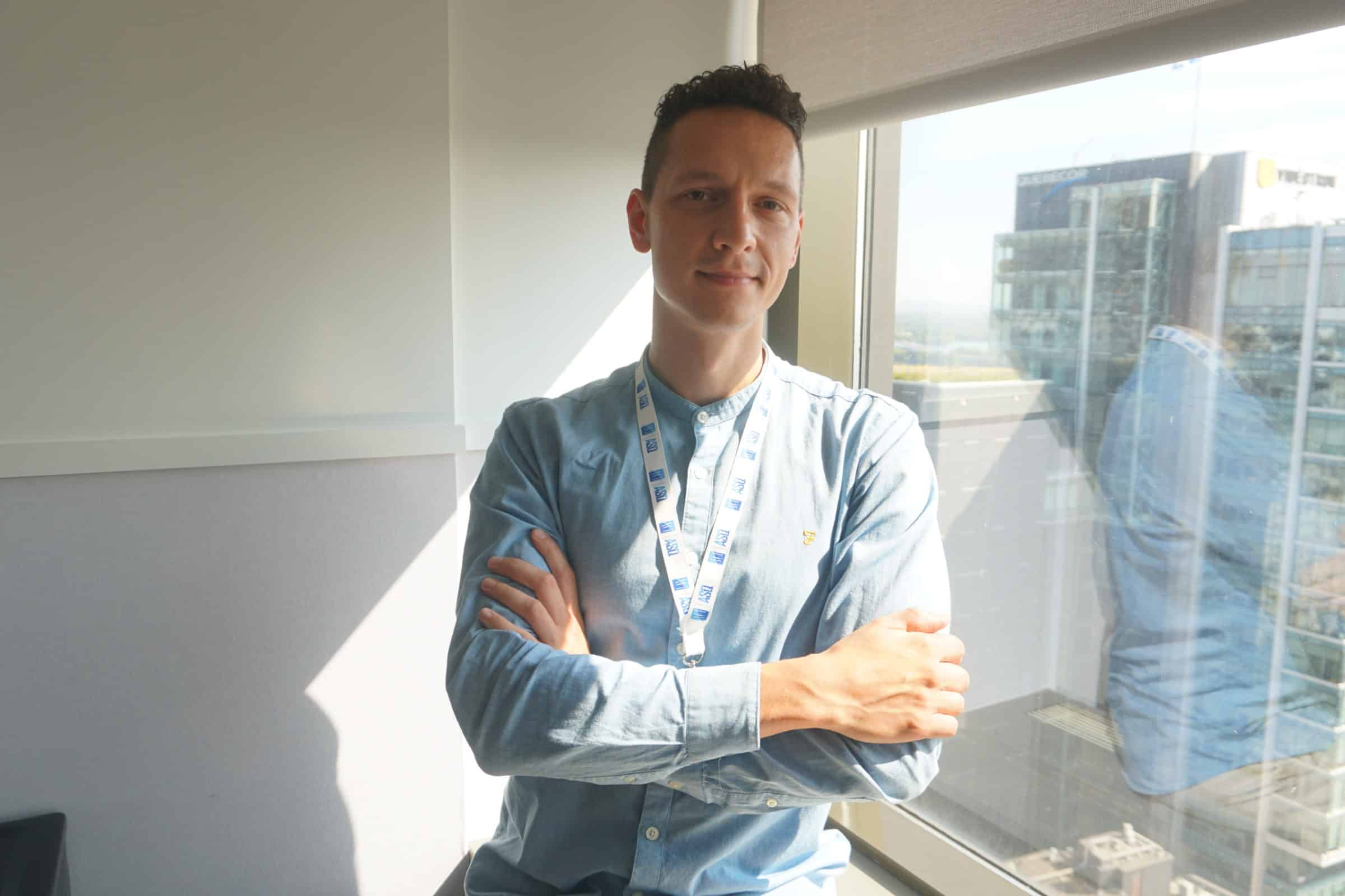 A man wearing a blue shirt and a lanyard stands with arms crossed by a sunny window in a modern office with city buildings visible outside.