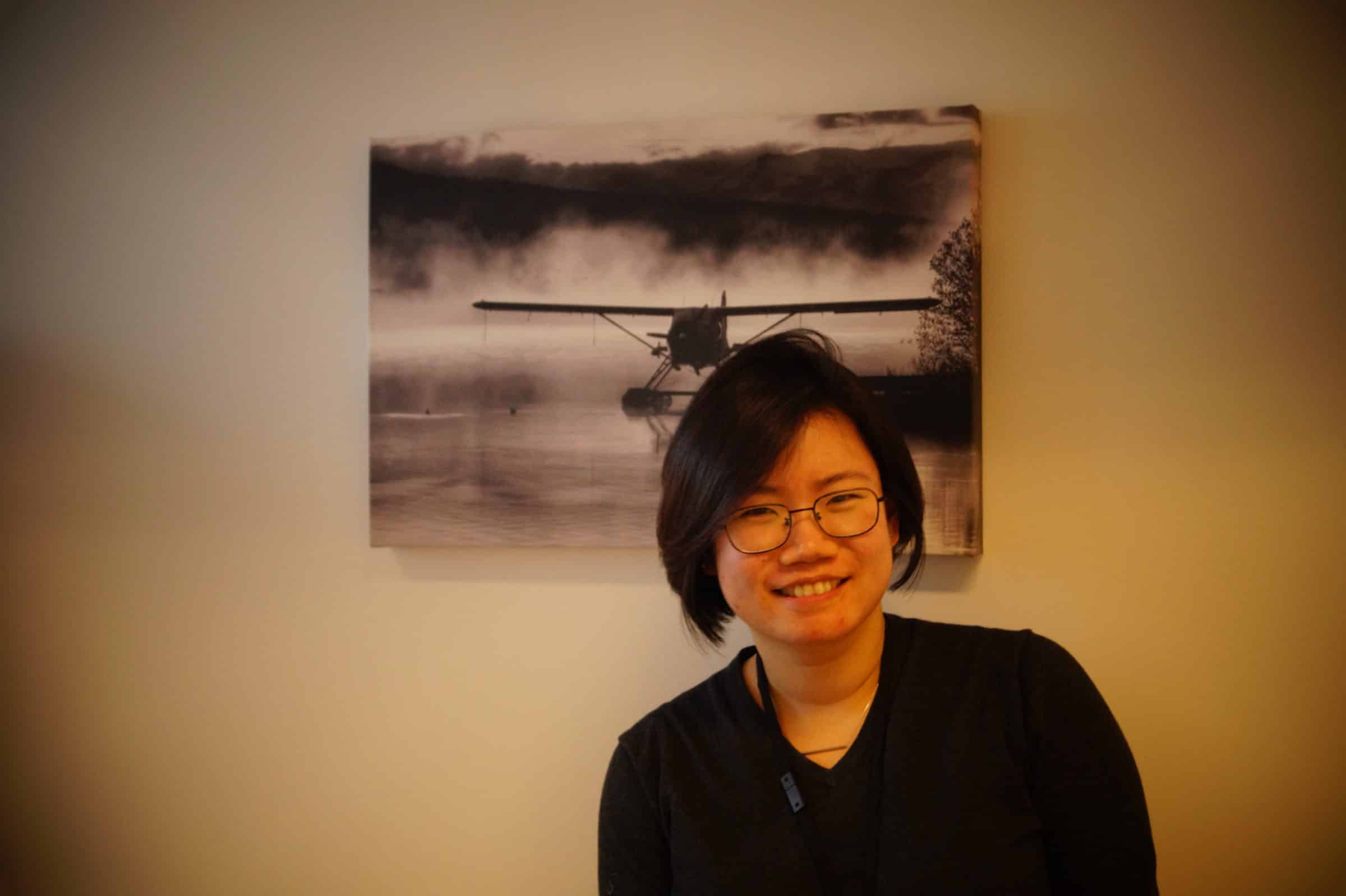A person with short dark hair and glasses smiles at the camera, standing in front of a wall with a black-and-white photo of a seaplane on a misty lake.