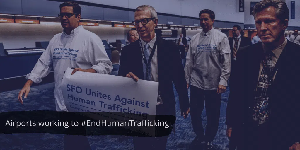 A group of people walk through an airport terminal, some wearing shirts and holding a sign that reads “SFO Unites Against Human Trafficking.” Text overlay says, “Airports working to #EndHumanTrafficking.”.