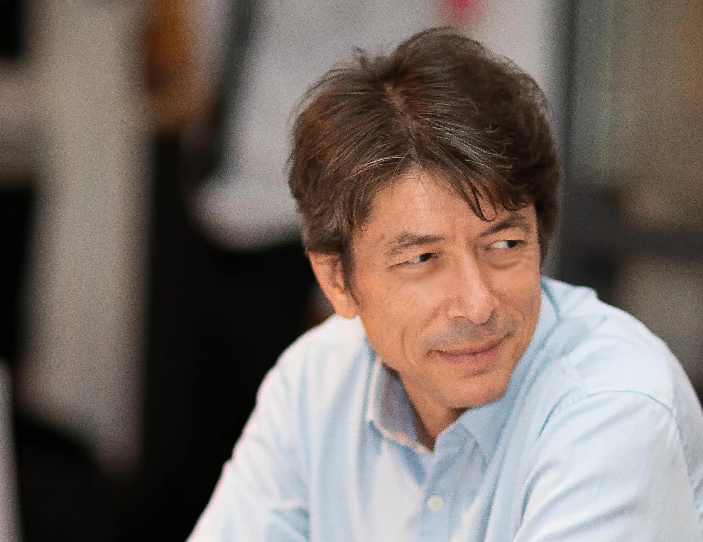A middle-aged man with brown hair wearing a light blue collared shirt is sitting indoors, slightly smiling and looking to his right. The background is softly blurred.