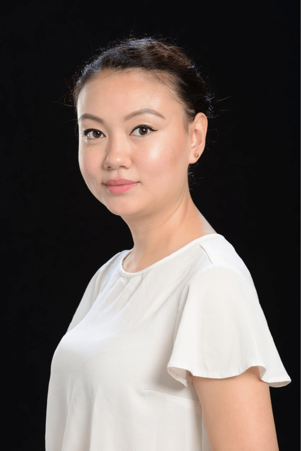 A woman with light skin and dark hair styled in a neat bun is wearing a white blouse and looking at the camera with a neutral expression against a plain black background.