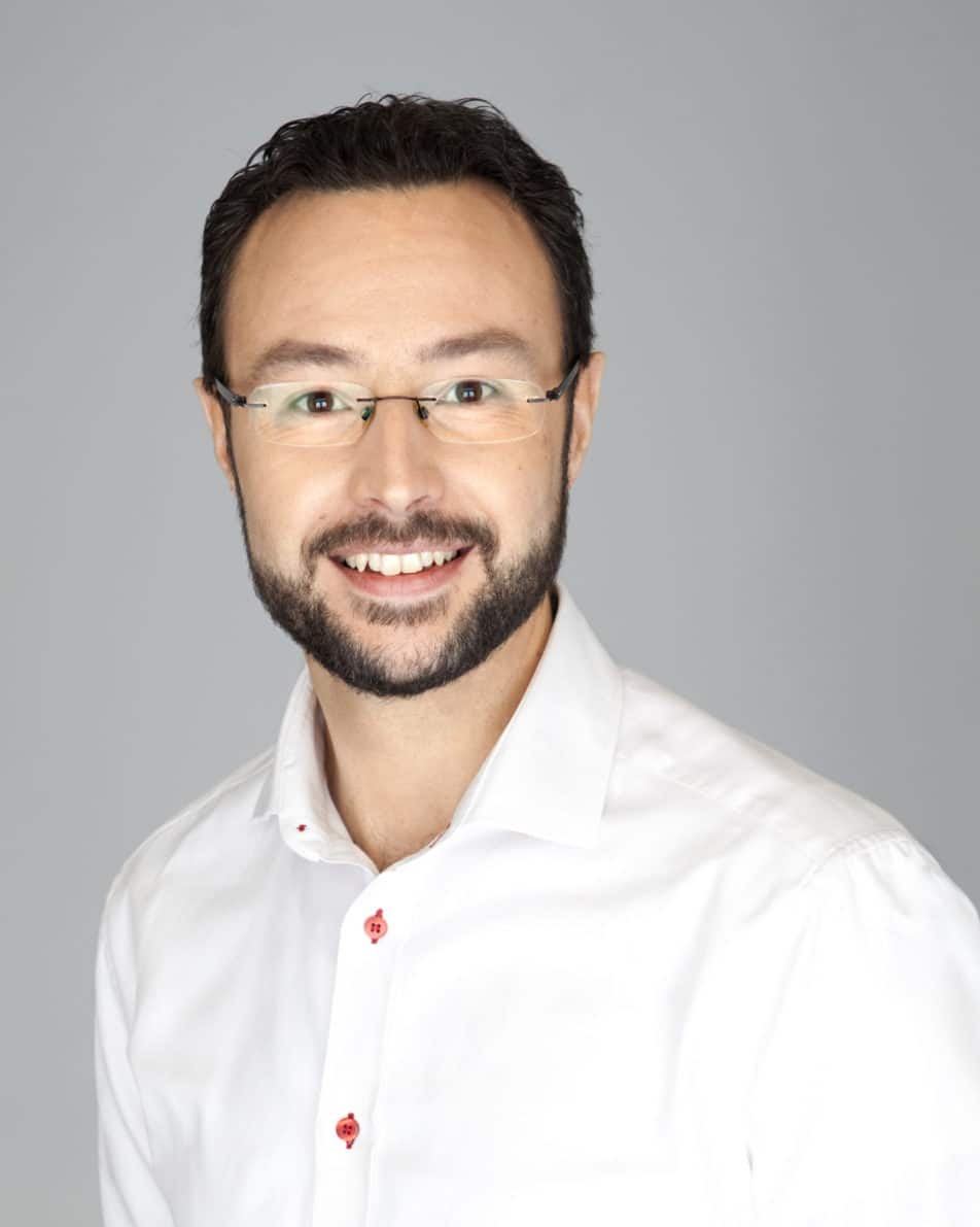 A man with short dark hair, a beard, and glasses, smiling at the camera. He is wearing a white shirt with red buttons, against a plain light gray background.