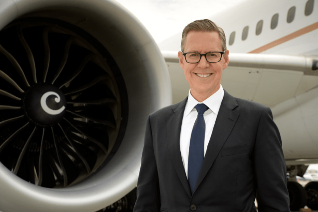 A man in a suit and tie smiles while standing in front of a large airplane engine, with the airplane’s fuselage visible in the background.