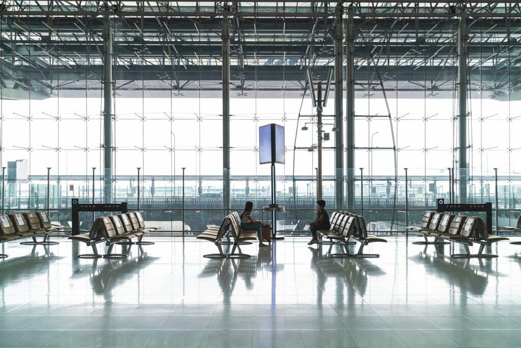 Two people sit apart on benches in a spacious, modern airport terminal with large glass windows, metal beams, and reflections on the shiny floor. The terminal appears bright and mostly empty.