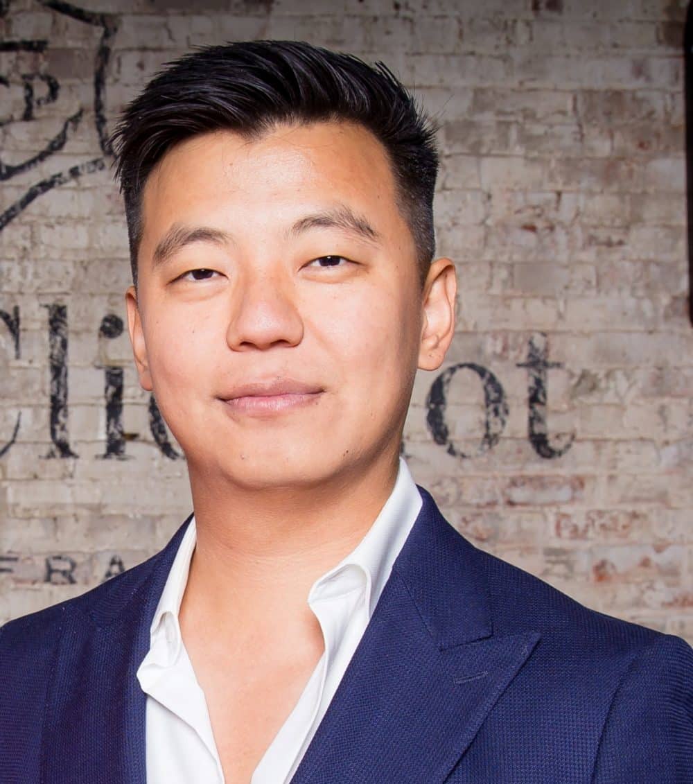 A man with short black hair wearing a navy blue suit jacket and white shirt stands in front of a brick wall with faded text. He is looking at the camera with a slight smile.