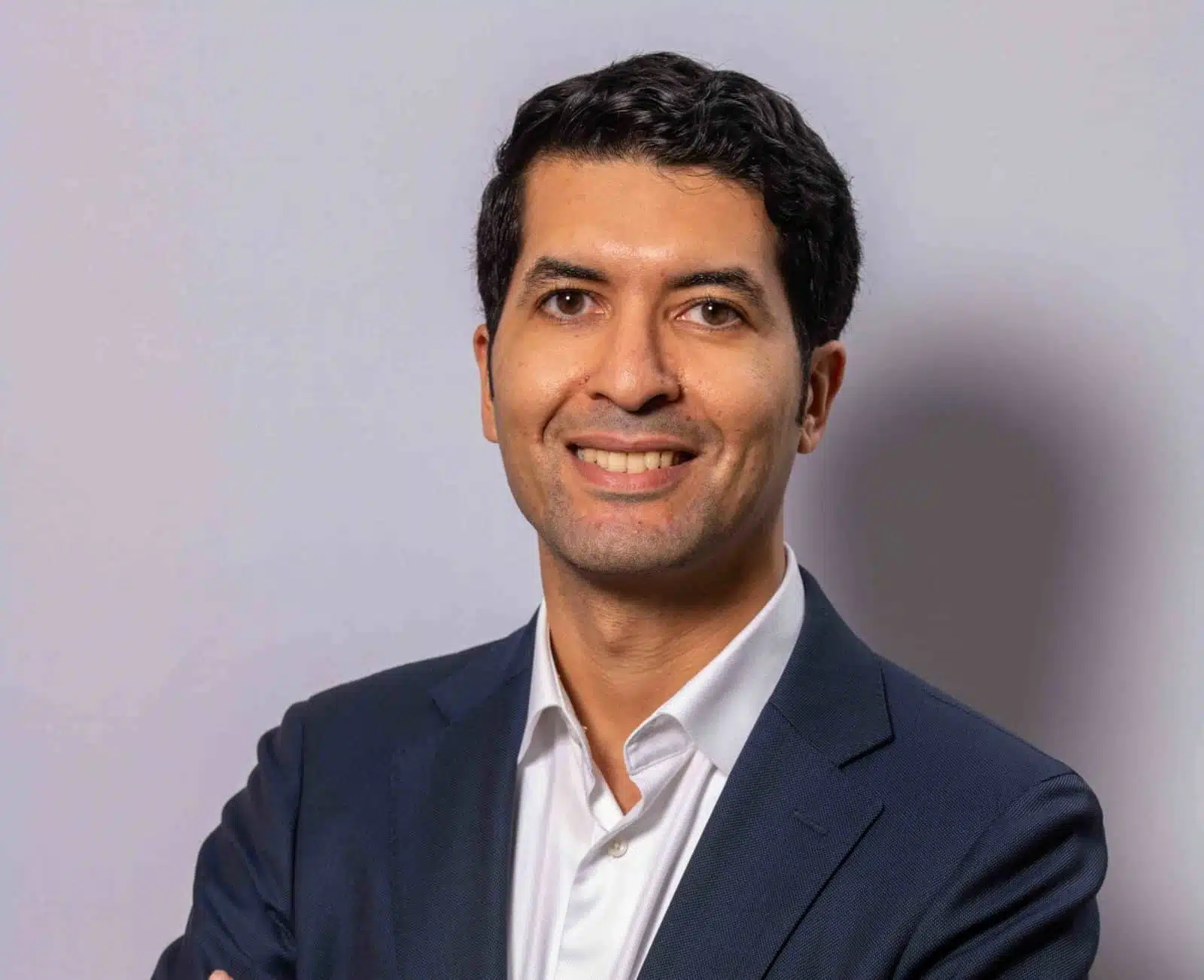 A man with short dark hair, wearing a dark suit jacket and white shirt, smiles confidently with arms crossed against a plain light gray background.