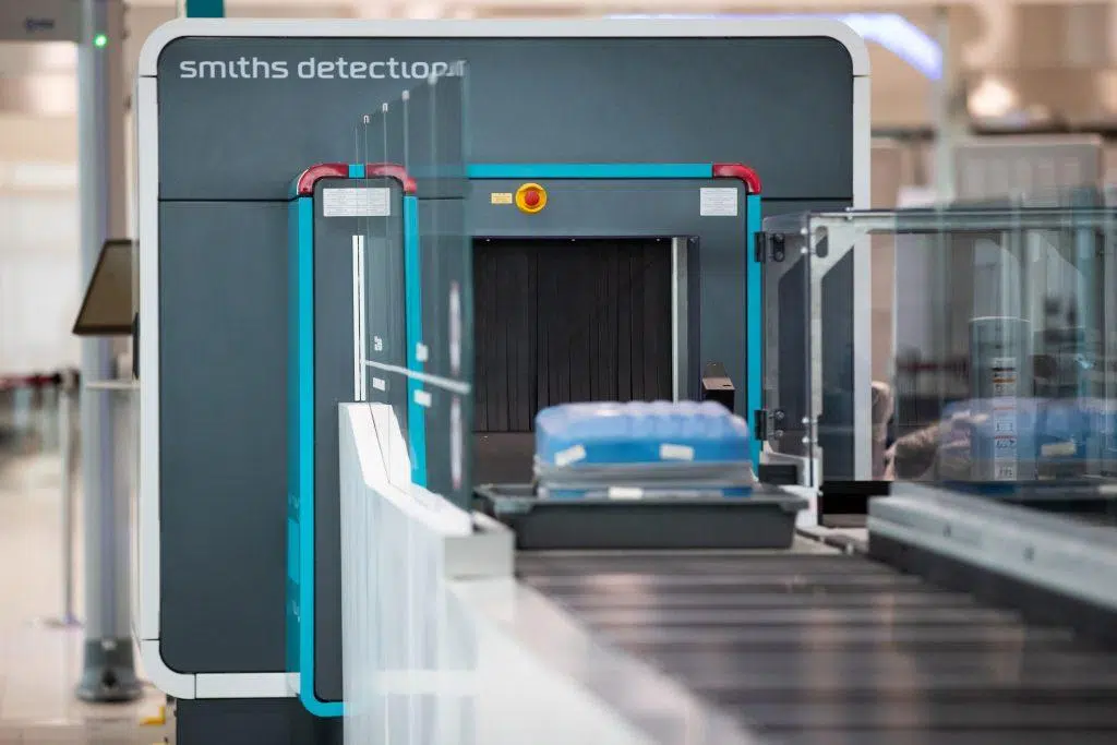 Airport security scanner with a conveyor belt carrying a blue plastic tray, used for screening luggage and personal items. The machine is labeled “Smiths Detection.”.