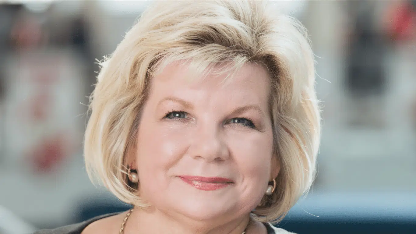 A middle-aged woman with short blonde hair and light makeup is smiling gently at the camera. She is wearing a gold necklace and earrings. The background is softly blurred.