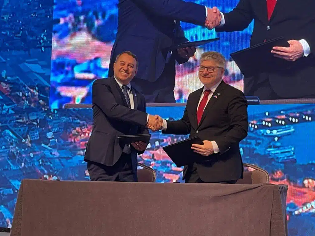 Two men in suits stand behind a table, smiling and shaking hands while holding folders. A large screen behind them displays a close-up of their handshake. The setting appears to be a formal event or agreement signing.