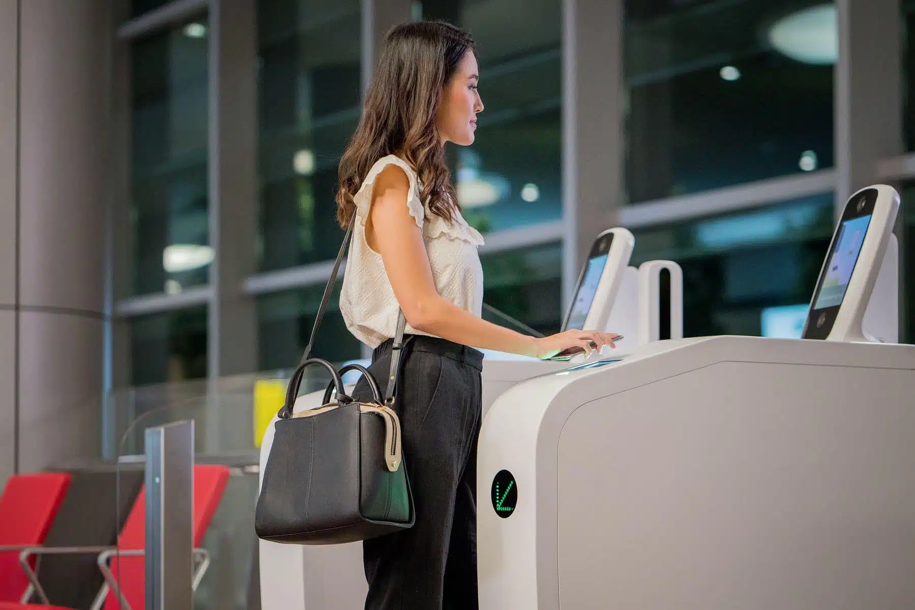 A woman stands at a self-service kiosk in an indoor setting, using a touchscreen with one hand while holding a black handbag. The background includes large windows and red chairs.