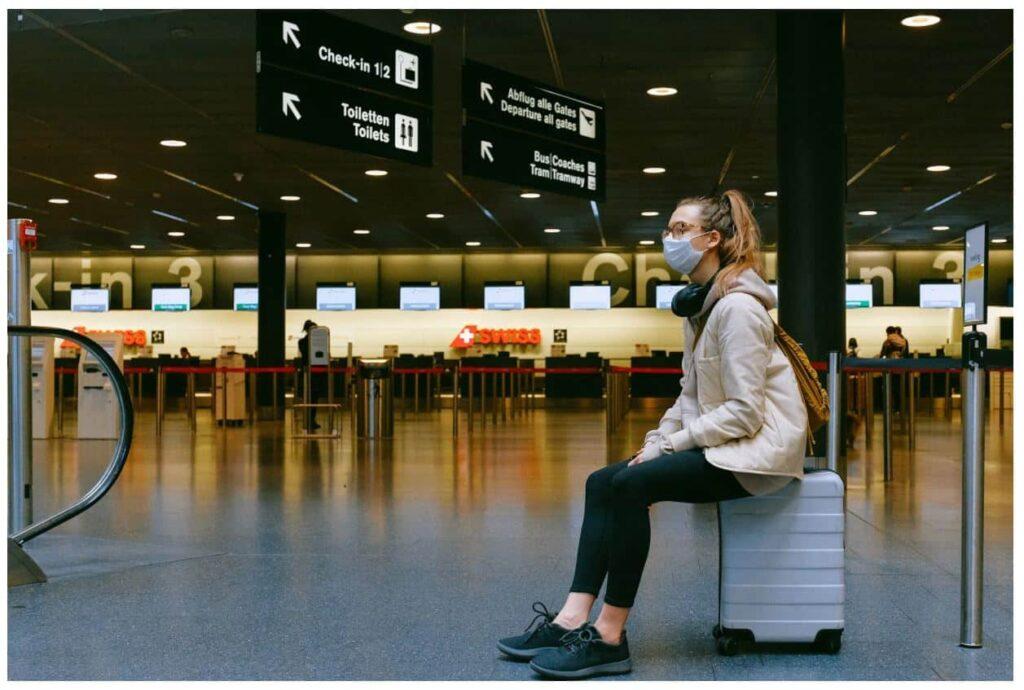 A woman wearing a face mask sits on a suitcase in an airport terminal, with check-in counters and overhead signs visible in the background.
