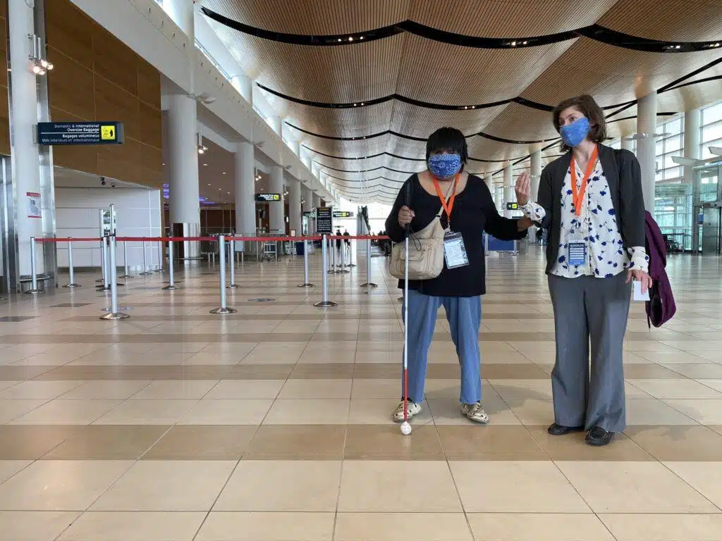 Two women wearing face masks stand in a spacious, modern airport terminal. One holds a cane and an ID badge, while the other waves and holds documents. The terminal appears mostly empty.